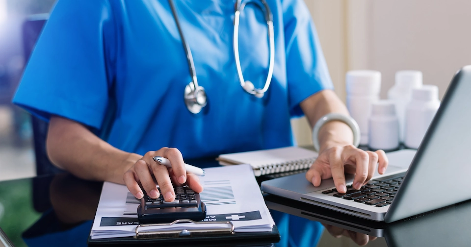 Healthcare worker with a stethoscope around the neck sitting at a laptop while entering data using a calculator