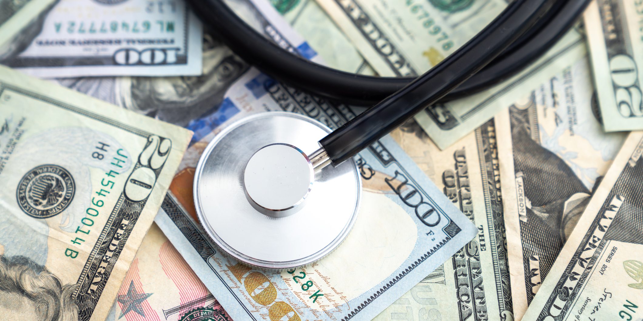 Doctor or nurse stethoscope medical device or equipment with metal parts and black tubing laying on a pile of united states currency in large bills or cash covering the entire surface.